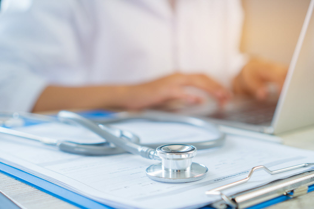 Provider on a laptop, stethoscope on a clipboard in front.